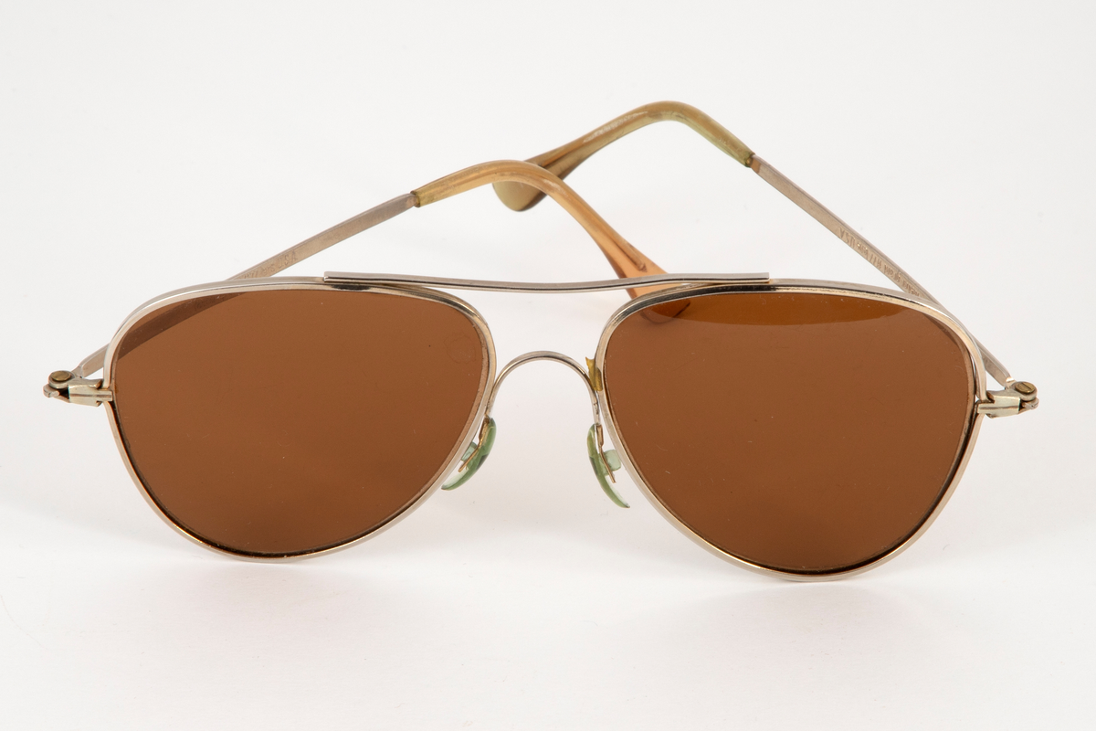 Solbriller med dråpeformede glass og smal metallinnfatning med en stang øverst, som forbinder glassinnfatningene. Smale, hengslede metallstenger som er plastbesatt på endene. Bevegelige neseputer i plast.