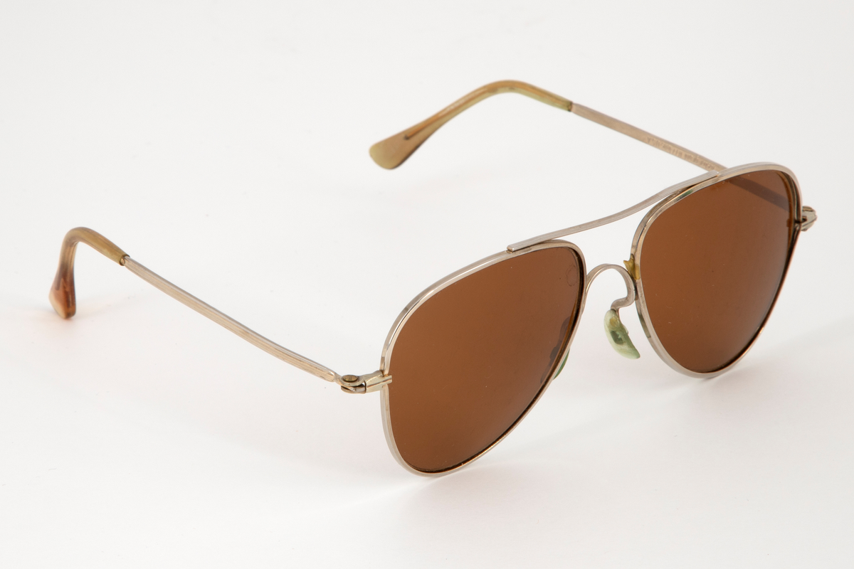 Solbriller med dråpeformede glass og smal metallinnfatning med en stang øverst, som forbinder glassinnfatningene. Smale, hengslede metallstenger som er plastbesatt på endene. Bevegelige neseputer i plast.