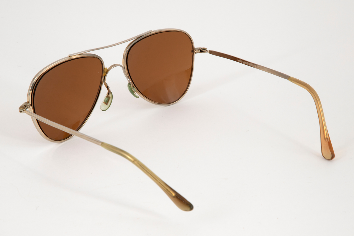 Solbriller med dråpeformede glass og smal metallinnfatning med en stang øverst, som forbinder glassinnfatningene. Smale, hengslede metallstenger som er plastbesatt på endene. Bevegelige neseputer i plast.