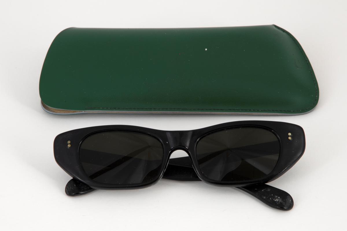 Solbriller med smale, ovale plastglass og Cat-Eye-formet, svart plastinnfatning. Hengslede stenger i svart plast med dråpeformet avslutning. Grønn rektangulært etui i skinn eller skinnimitiat med åpning i den ene kortsiden.