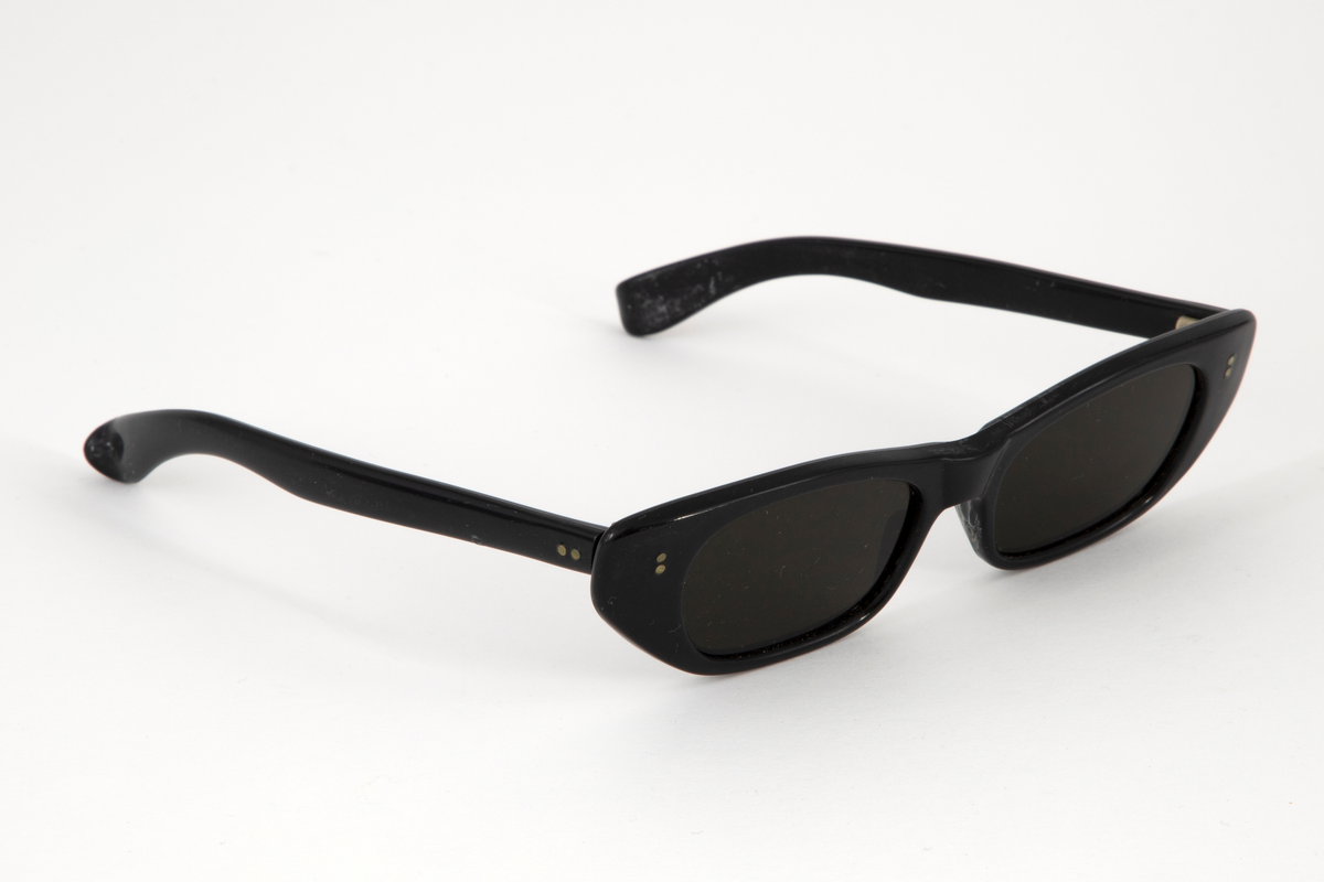 Solbriller med smale, ovale plastglass og Cat-Eye-formet, svart plastinnfatning. Hengslede stenger i svart plast med dråpeformet avslutning. Grønn rektangulært etui i skinn eller skinnimitiat med åpning i den ene kortsiden.