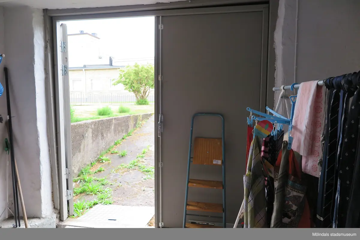 En öppen dörr (av två) från ett vitmålat förråd/garage i källarplan ut till trädgården, Krokslättsgatan 18 i Mölndal 2023-06-21. På höger sida om dörren står en trappstege samt en stång med kläder. Fotodokumentation av ett friliggande bostadshus/villa byggt 1932 med fyra (4) våningsplan. Byggnaden har tvättstuga i källaren samt torkvind på övervåningen. Boarea: 140 kvm, totalarea: 1004 kvm. Villan är ombyggd och fördelad på fyra (4) hyreslägenheter.
Relaterade motiv: 2024_1415 - 1541.