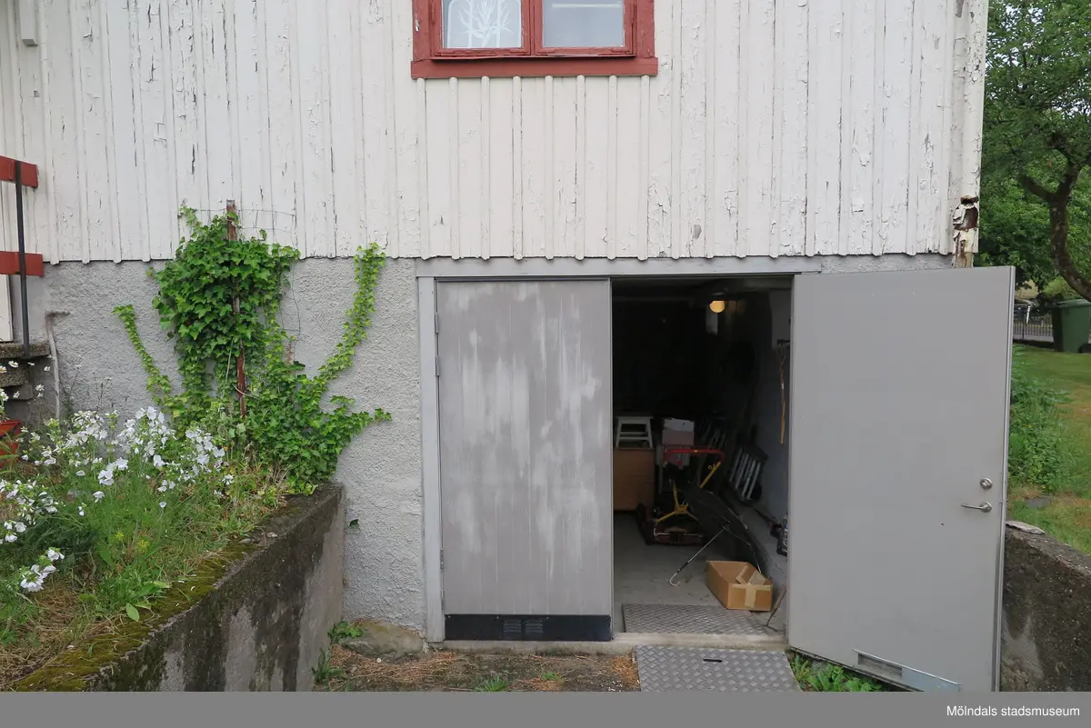 Ingång från trädgården till förråd/garage i källarplan (se 2024_1530), Krokslättsgatan 18 i Mölndal 2023-06-21. Till vänster ses växlighet. Rakt fram ses en del av den vita husfasaden samt en (av två) öppen dörr till förrådet. Fotodokumentation av ett friliggande bostadshus/villa byggt 1932 med fyra (4) våningsplan. Byggnaden har tvättstuga i källaren samt torkvind på övervåningen. Boarea: 140 kvm, totalarea: 1004 kvm. Villan är ombyggd och fördelad på fyra (4) hyreslägenheter.
Relaterade motiv: 2024_1415 - 1541.