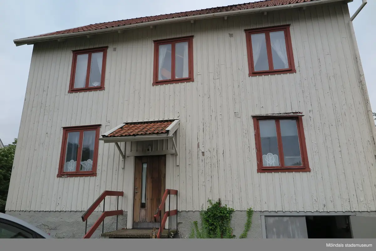 Bostadshus med vit träfasad, huvudentré med trappa, fem fönster och källarplan, Krokslättsgatan 18 i Mölndal 2023-06-21. Lägenheternas fönster är målade i roströd färg. Till höger ses förråd/garage i källarplan. Fotodokumentation av ett friliggande bostadshus/villa byggt 1932 med fyra (4) våningsplan. Byggnaden har tvättstuga i källaren samt torkvind på övervåningen. Boarea: 140 kvm, totalarea: 1004 kvm. Villan är ombyggd och fördelad på fyra (4) hyreslägenheter.
Relaterade motiv: 2024_1415 - 1541.