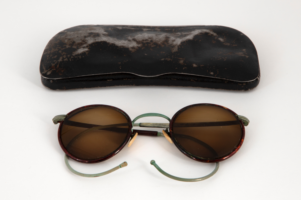 Solbriller med runde, lett dråpeformede, brunfargede glass innfattet i en smal metallramme belagt med brun plast, antagelig bakelitt. Smale stenger belagt med samme materiale og grønne, sterk svungne, bevegelige ender. Grønn nesebro og neseputer av hvit plast. Avlang brilleetui i svartlakkert metall med hengslet lokk. Etuiet er innvendig kledd med rød fløyel.