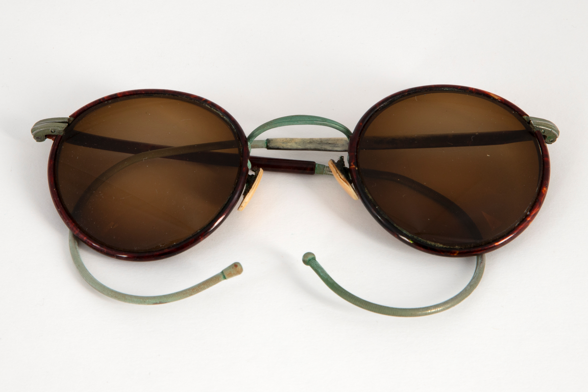 Solbriller med runde, lett dråpeformede, brunfargede glass innfattet i en smal metallramme belagt med brun plast, antagelig bakelitt. Smale stenger belagt med samme materiale og grønne, sterk svungne, bevegelige ender. Grønn nesebro og neseputer av hvit plast. Avlang brilleetui i svartlakkert metall med hengslet lokk. Etuiet er innvendig kledd med rød fløyel.