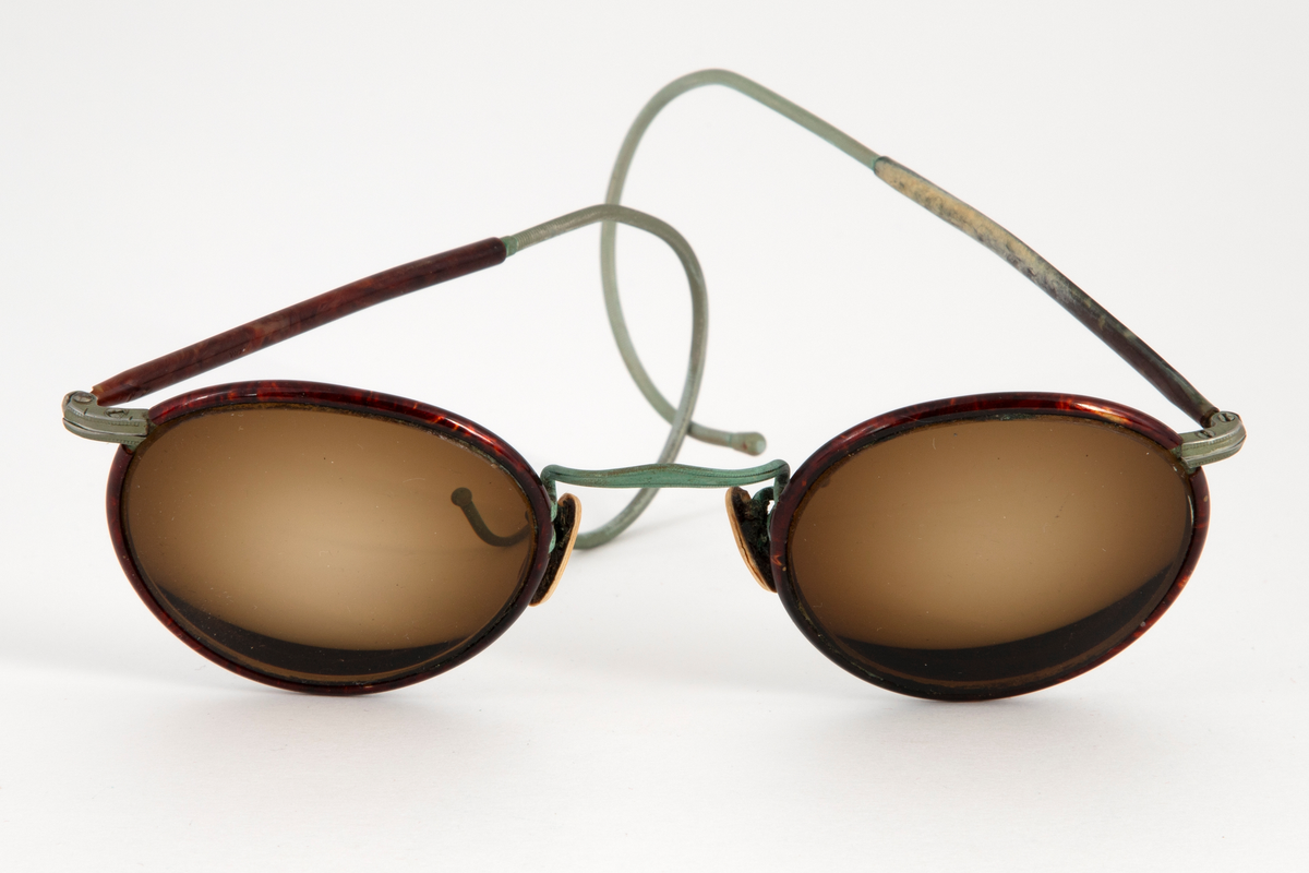 Solbriller med runde, lett dråpeformede, brunfargede glass innfattet i en smal metallramme belagt med brun plast, antagelig bakelitt. Smale stenger belagt med samme materiale og grønne, sterk svungne, bevegelige ender. Grønn nesebro og neseputer av hvit plast. Avlang brilleetui i svartlakkert metall med hengslet lokk. Etuiet er innvendig kledd med rød fløyel.