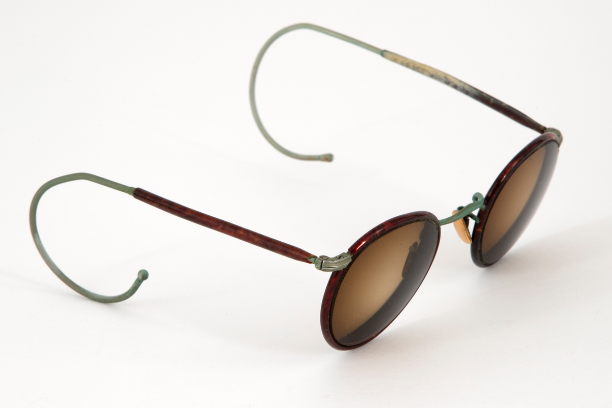 Solbriller med runde, lett dråpeformede, brunfargede glass innfattet i en smal metallramme belagt med brun plast, antagelig bakelitt. Smale stenger belagt med samme materiale og grønne, sterk svungne, bevegelige ender. Grønn nesebro og neseputer av hvit plast. Avlang brilleetui i svartlakkert metall med hengslet lokk. Etuiet er innvendig kledd med rød fløyel.