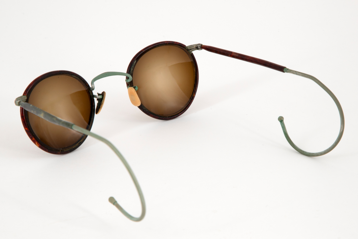 Solbriller med runde, lett dråpeformede, brunfargede glass innfattet i en smal metallramme belagt med brun plast, antagelig bakelitt. Smale stenger belagt med samme materiale og grønne, sterk svungne, bevegelige ender. Grønn nesebro og neseputer av hvit plast. Avlang brilleetui i svartlakkert metall med hengslet lokk. Etuiet er innvendig kledd med rød fløyel.