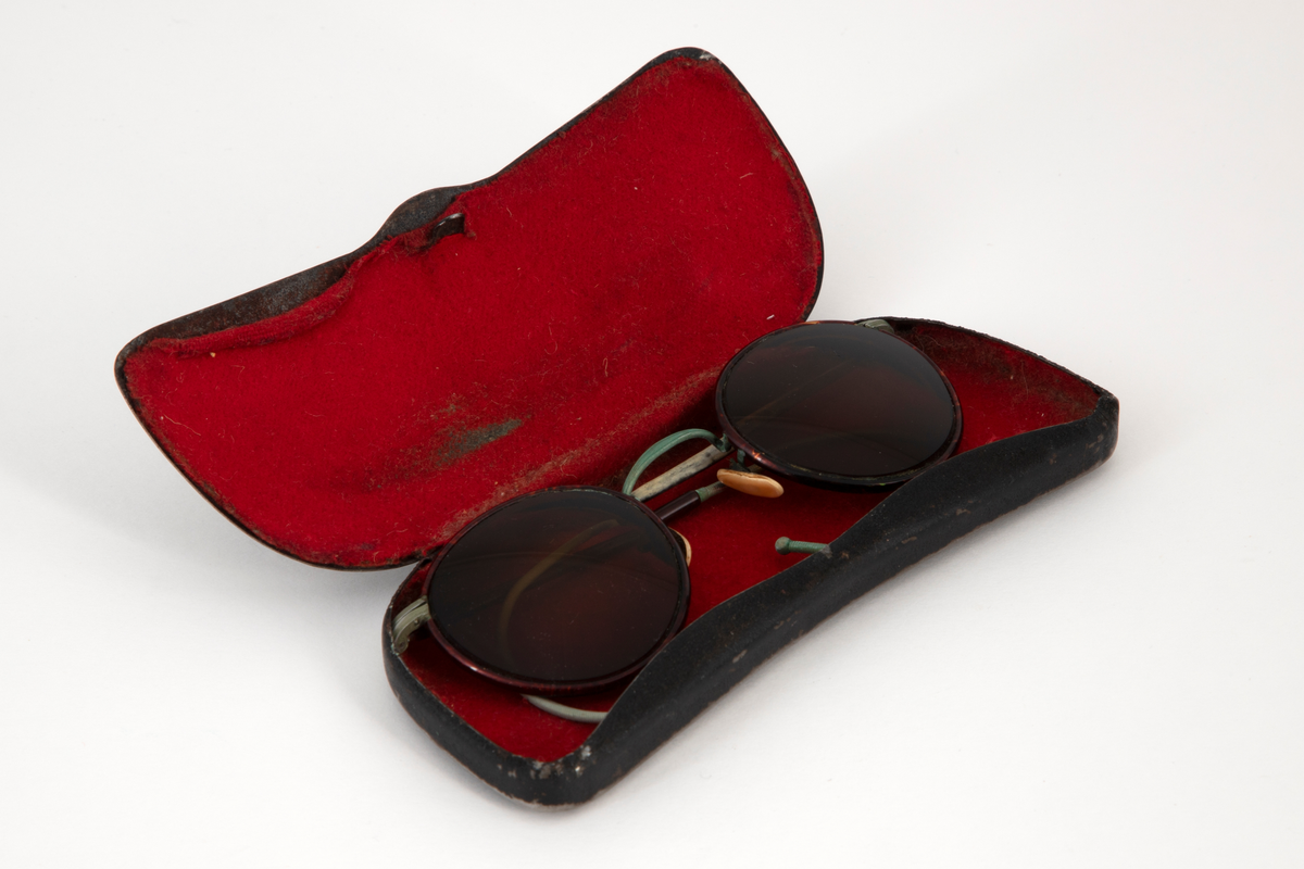 Solbriller med runde, lett dråpeformede, brunfargede glass innfattet i en smal metallramme belagt med brun plast, antagelig bakelitt. Smale stenger belagt med samme materiale og grønne, sterk svungne, bevegelige ender. Grønn nesebro og neseputer av hvit plast. Avlang brilleetui i svartlakkert metall med hengslet lokk. Etuiet er innvendig kledd med rød fløyel.
