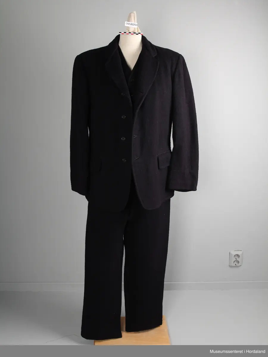 Herredress av vadmål beståande av bukse, jakke og dress:

1) Langbukse av mørkt blått vadmålstoff. Gylven er midt på framfor og har knappar og ei hekte. Buksa har ei lomme på kvar side. På baksida er det ein splitt i ryggen og eit spensel.

2) Jakke av mørkt blått vadmålstoff. Den har slag, opning framfor med fire knappar og knappehol, og dessutan to knappar nedst på ermane. Innvendig er jakka fôra med eit mørkt, rutete stoff. Jakka har to klafflommer med fôra klaffar i same rutete stoff.

3) Vest av mørkt blått vadmålstoff. Den har seks knappar med knappehol i opninga framfor, og dessutan to lommer på den eine sida og ei lomme på den andre sida. Bakstykket er av grått bomullsstoff med spennsel. Vesten er fôra.
