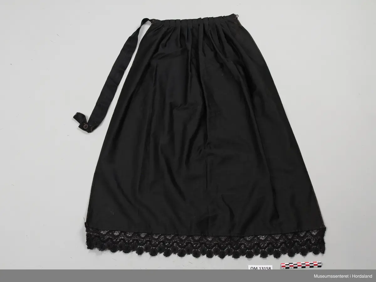 Forkle av sateng. Det har brunt bomull i renning og svart silke i innslag. Bred blondekant nederst påsydd med maskin. Forkleet er lagt i legg oppe i livet, uten linning, men med støttebånd på baksiden. Her er også et silkebånd som kan festes med knapp i en hempe laget av knappehullssting. Enden av silkebåndet er til dels revet av og henger i en tynn tråd. Det er en rift som er sydd igjen under hempen. Sidene har jarekanter.