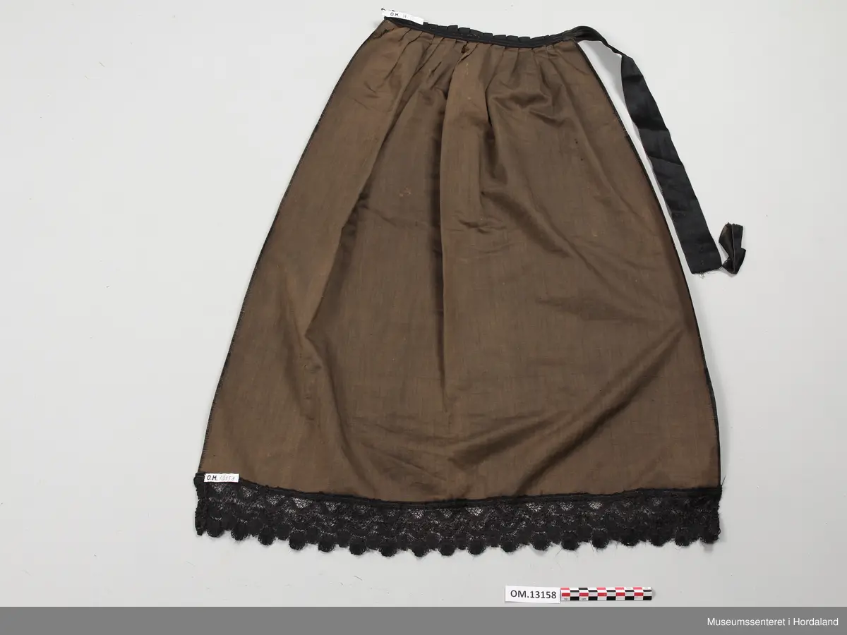 Forkle av sateng. Det har brunt bomull i renning og svart silke i innslag. Bred blondekant nederst påsydd med maskin. Forkleet er lagt i legg oppe i livet, uten linning, men med støttebånd på baksiden. Her er også et silkebånd som kan festes med knapp i en hempe laget av knappehullssting. Enden av silkebåndet er til dels revet av og henger i en tynn tråd. Det er en rift som er sydd igjen under hempen. Sidene har jarekanter.