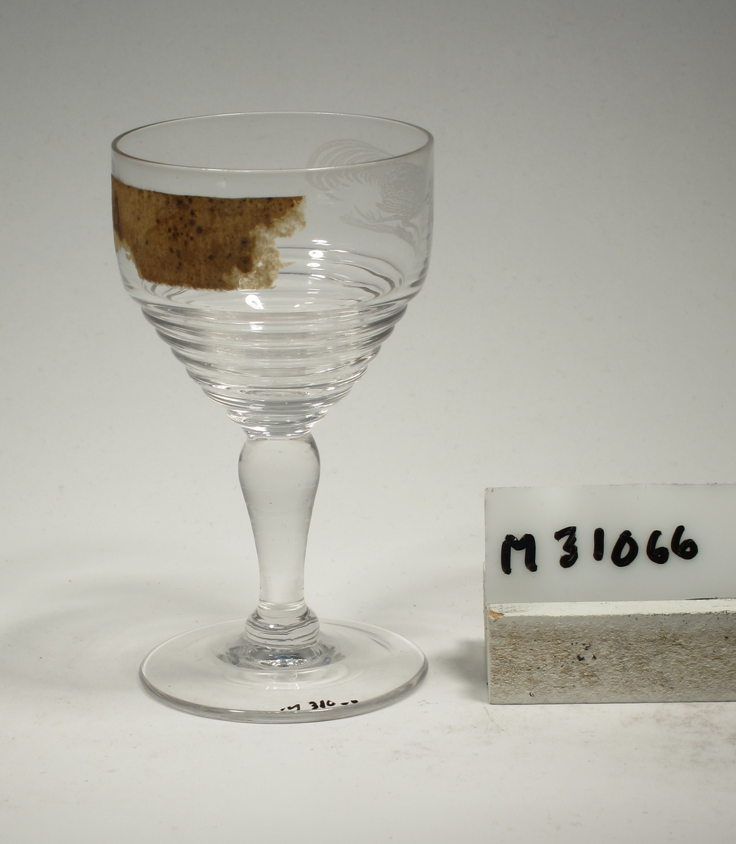 Formgivare: Ingrid Atterberg. Cocktailglas på ben och fot. Ovoid kupa med horisontella vulster på nedre delen. Etsad tupp på övre delen. Pelarben.