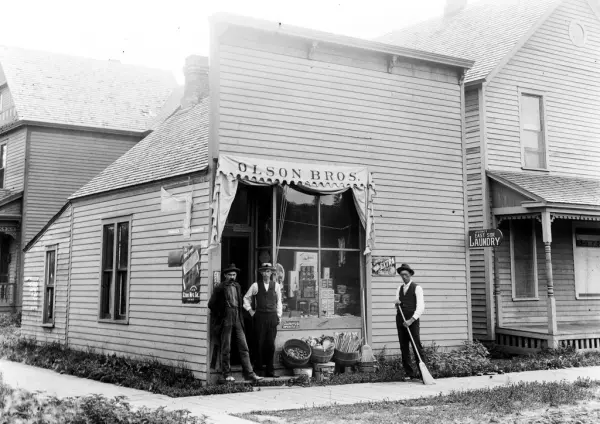 Butikk "Olson Bros", tre menn utenfor, - den ene med feiekos