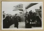 Bilden visar en amiral och sjöofficerare som har tagit uppställning ombord på pansarskeppet Sverige och hälsar kung Gustav V. I bakgrunden syns ett flertal fotografer som dokumenterar händelsen.