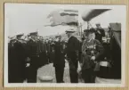 Bilden visar en amiral och sjöofficerare som har tagit uppställning ombord på pansarskeppet Sverige och hälsar kung Gustav V. I bakgrunden syns ett flertal fotografer som dokumenterar händelsen. I förgrund står en general med många ordnar och medaljer.