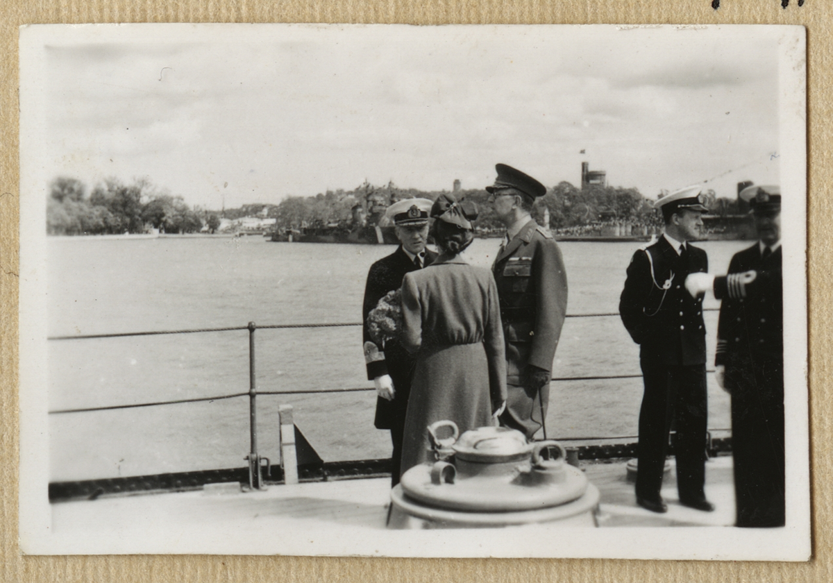 Bilden visar kronprinsessan Louise och kronprins Gustav i samtal med en amiral ombord på pansarskepp Sverige. I bakgrunden syns Kastellholmen där ett större antal örlogsfartyg ligger förtöjd.