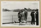 Bilden visar kronprinsessan Louise och kronprins Gustav i samtal med en amiral ombord på pansarskepp Sverige. I bakgrunden syns Kastellholmen där ett större antal örlogsfartyg ligger förtöjd.
