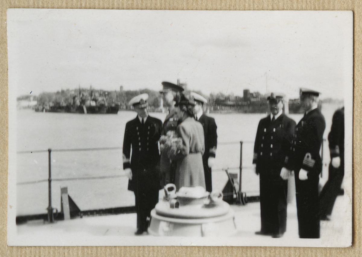 Bilden visar kronprinsessan Louise och kronprins Gustav i samtal med en amiral ombord på pansarskepp Sverige. I bakgrunden syns Kastellholmen där ett större antal örlogsfartyg ligger förtöjd.