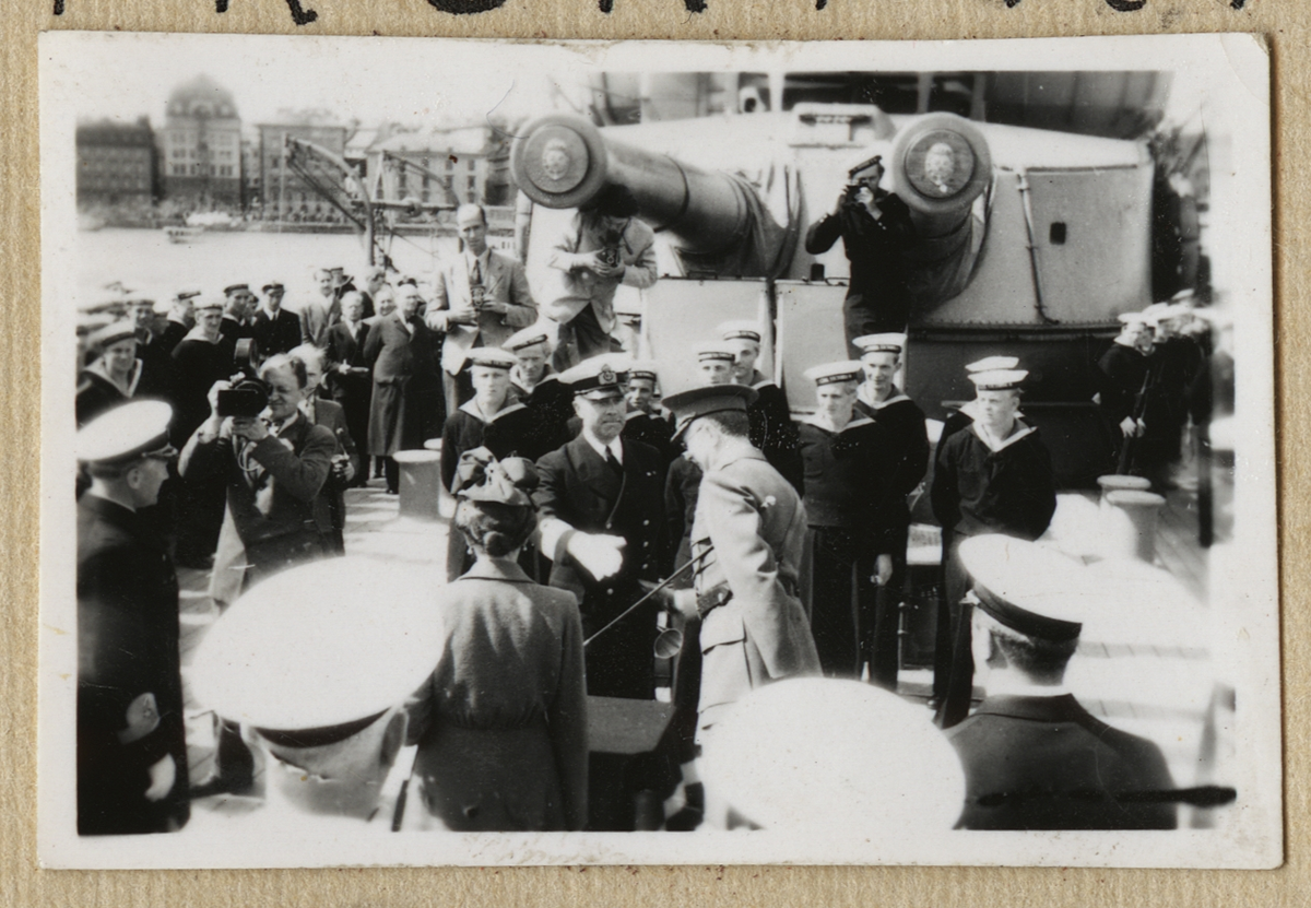 Bilden visar kronprinsessan Louise och kronprins Gustav som hälsas välkommen ombord på pansarskepp Sverige. I bakgrunden syns en fotograf och en kameraman som dokumenterar händelsen.
