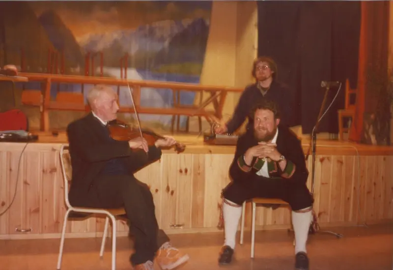 Anton Svendsen Myrvoll spiller fele mens Svein Aakerholt lytter. Øystein Gjerde står i bakgrunnen med lydopptaker. Bildet er tatt i forbindelse med et arrangement på Vassås i Hof i mai 1980, og opptaket er i dag en del av Gjerde-samlingen, og er åpent tilgjengelig via Vestfoldarkivets webside.