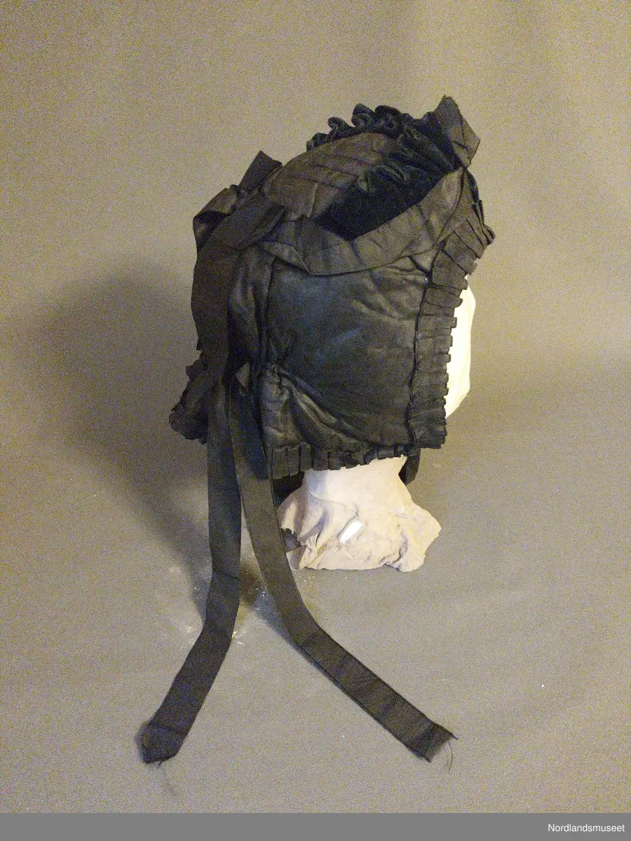 Svart kyse i silke (?), med knyttebånd til å knytte under haken og to sløyfer bakpå derfra pyntebånd henger ned. Mørkegrønnblå plysjdetaljer over pannen. Rynket bånd rund ansiktet og på nedkanten.