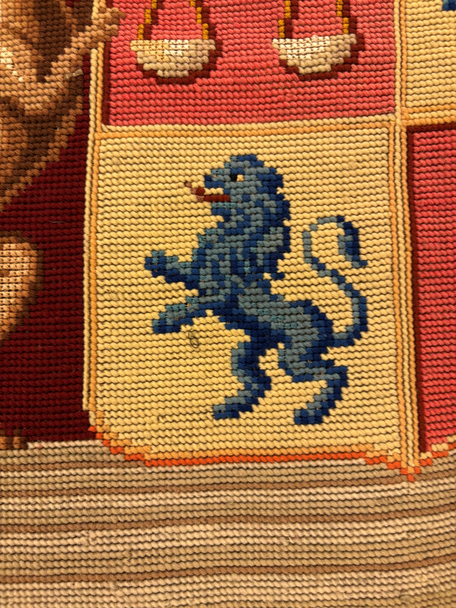 Matta eller broderad tapet, med yttäckande broderi, röd botten med blommönster i hörnen. I mitten en vapensköld (ätten Löwenhielm) med text i gult under. Snodd i silke och ylle runt om.