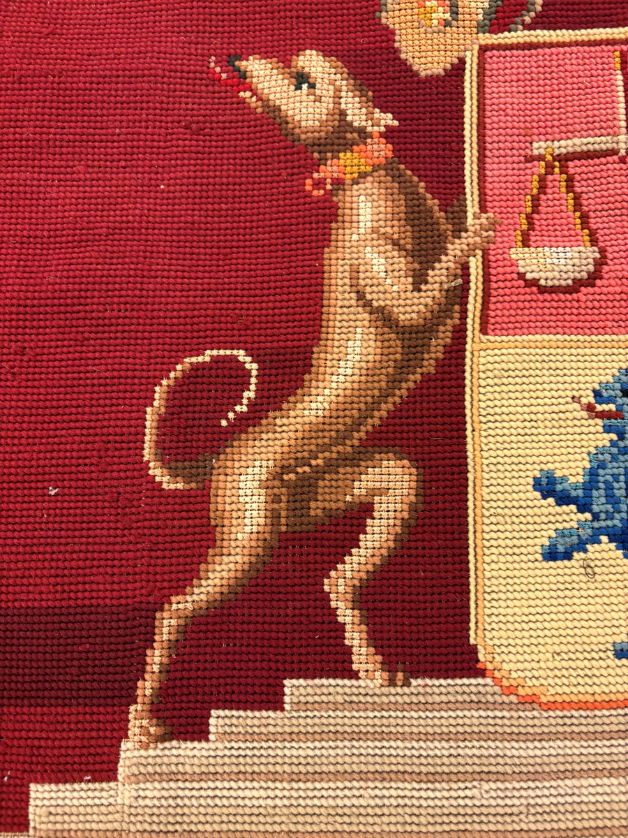 Matta eller broderad tapet, med yttäckande broderi, röd botten med blommönster i hörnen. I mitten en vapensköld (ätten Löwenhielm) med text i gult under. Snodd i silke och ylle runt om.