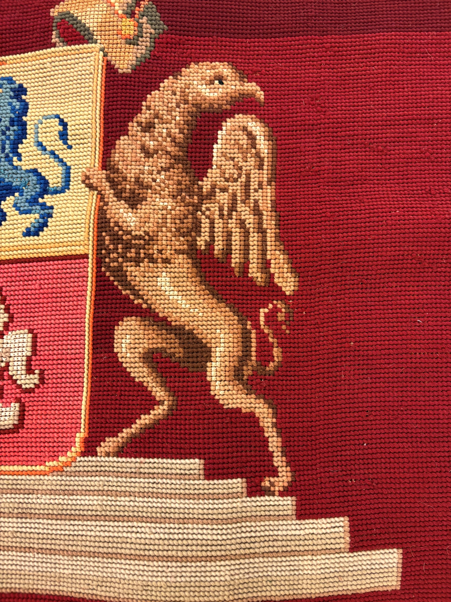 Matta eller broderad tapet, med yttäckande broderi, röd botten med blommönster i hörnen. I mitten en vapensköld (ätten Löwenhielm) med text i gult under. Snodd i silke och ylle runt om.