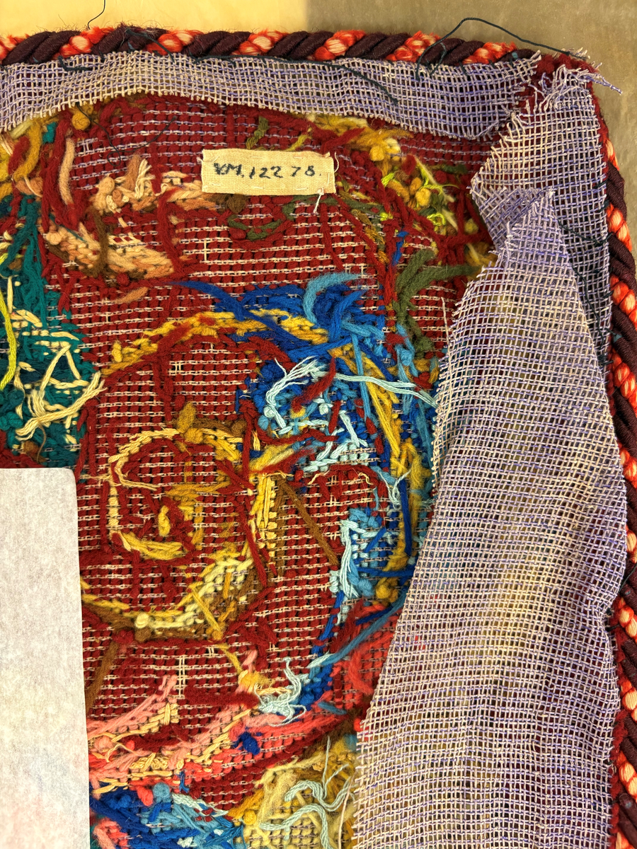 Matta eller broderad tapet, med yttäckande broderi, röd botten med blommönster i hörnen. I mitten en vapensköld (ätten Löwenhielm) med text i gult under. Snodd i silke och ylle runt om.