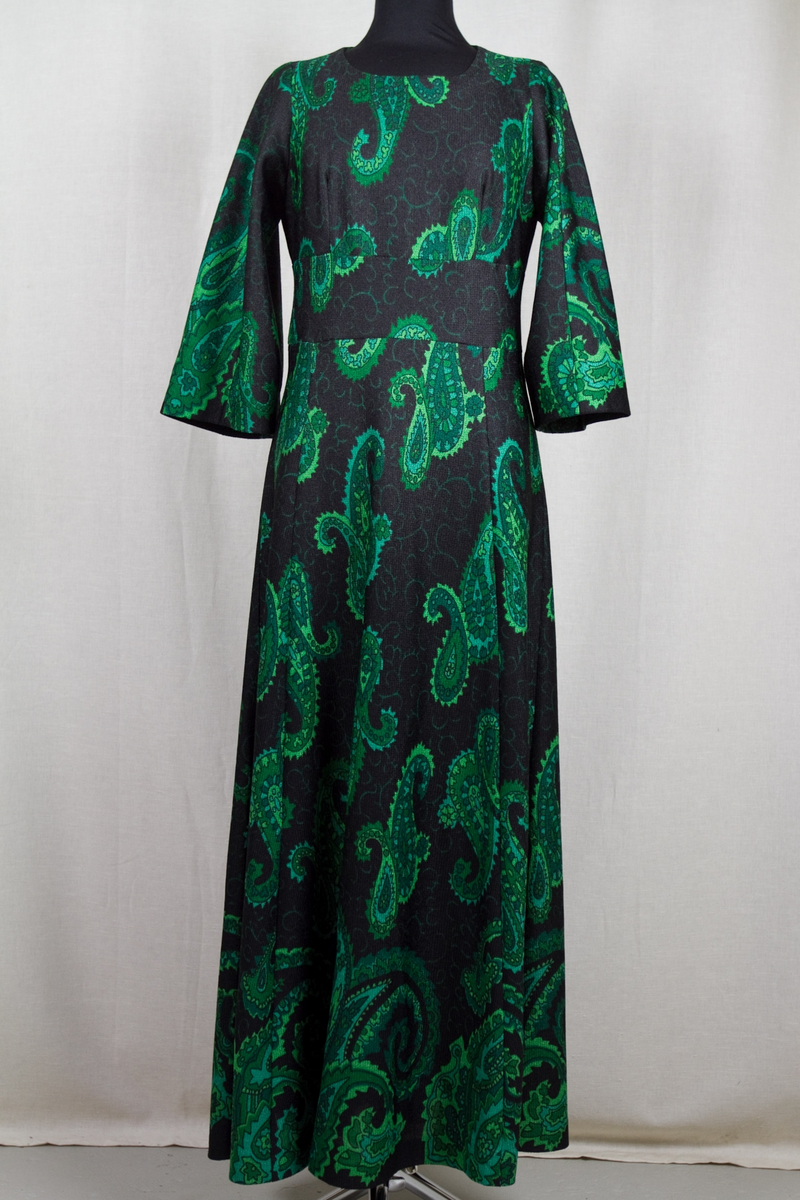 Långklänning, svart botten med persiskt tryckt mönster i olika gröna nyanser. Rund vid halsringning. Vid trekvartslång isydd tvåsömsärm. Delad under bysten med en 10 cm bred och slätt påsydd linning. Baktill en 59 cm lång dragkedja. Kjolen har 7 st utställda våder.