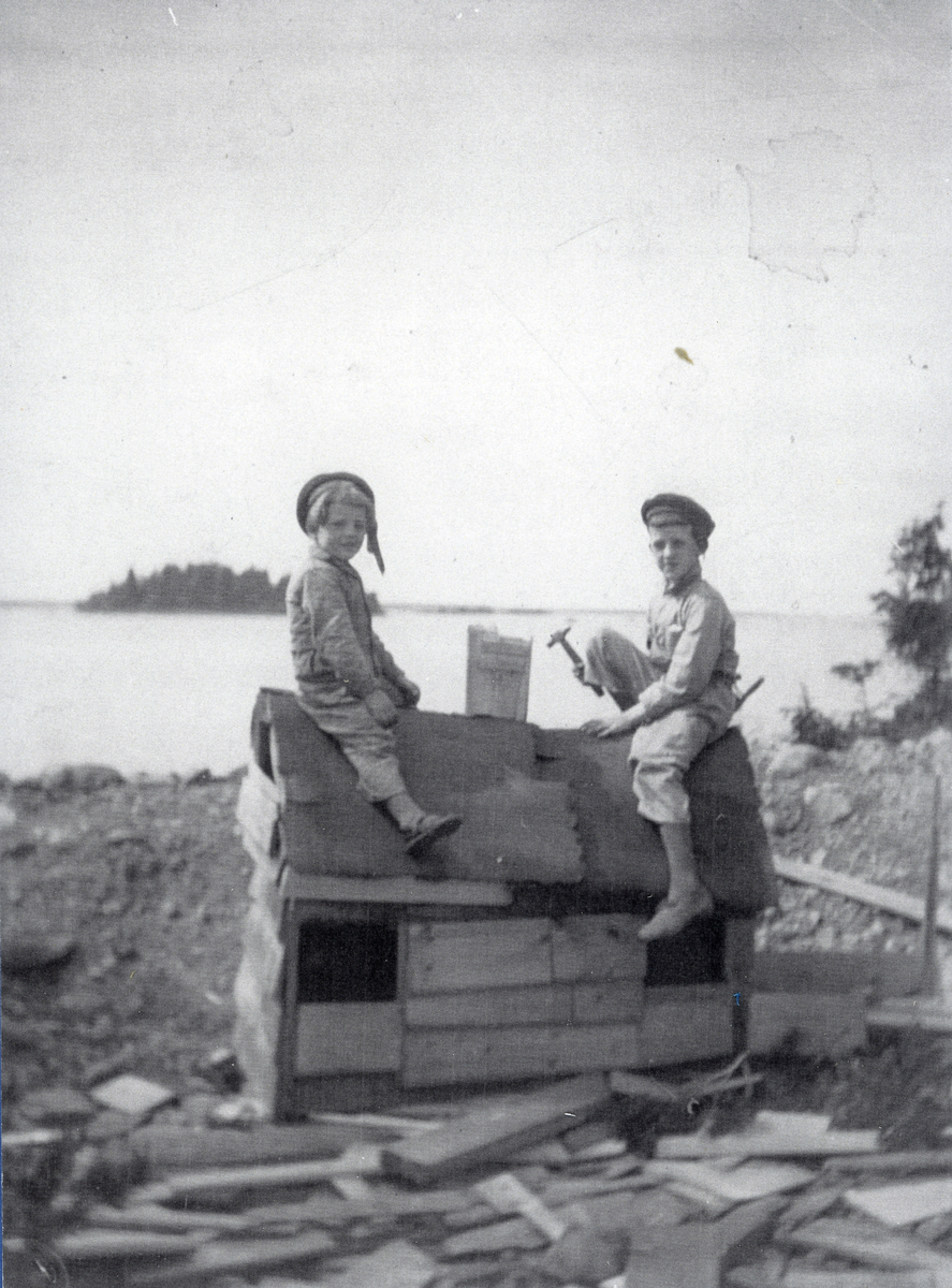 Olle och Sven bygger eget, sittandes på taknocken och med verktyg i handen. I bakgrunden syns en liten ö ute i vattnet. En liten kopia av stora "Limöhäll".
