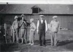 Arrendator Carl Eriksson och fiskare Axel Hillberg till höger samt tre personer till vänster, varav den i mitten håller i en valp, står framför en lång byggnad, 1929.