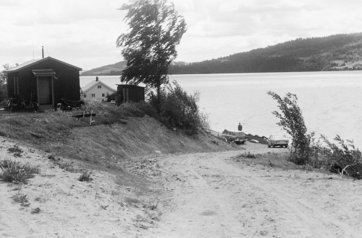 Til høyre på dette bildet ser vi en velteplass for tømmer i «bilbunter» ved Mjøs-stranda på Brøttum i Ringsaker kommune, altså i den nordøstre enden av innsjøen, bygd våren 1961. Utislagsplassen er bygd på to betongmurer - den ene er lav og befinner seg under vannspeilet ved strandkanten og er ikke synlig på dette fotografiet, den andre, som er noe høyere, er bygd langs en veg som akkurat her gikk parallelt med den nevnte strandkanten i tre-fire meters avstand. Vinkelrett på de to murene er det lagt ei slags rist - bestående av kraftige stålbjelker, Tømmerbilen skulle kjøres slik at den sto med lasten parallelt med denne rampa. Deretter skulle sjåføren felle ned stakene på denne sida av lasset slik at tømmeret - omsluttet av et par vaiergrimer - kunne falle ned på de skråttliggende stålbjekene og skli ut i vannet. Der ble det etter hvert hentet av en slepebåt som bukserte virket, enten til kjøpere i Mjøs-området, eller mot Minnesund i sørenden av innsjøen, der strømmen skulle bære det videre sørover i Vorma og Glomma. Da dette fotografiet ble tatt sto det to personbiler på den vegen tømmerbilene skulle kjøres på. Personbilene tilhørte antakelig fløtingsfunksjonærer som var på befaring. Begge ser ut til å være av merket Ford Taunus 17M, den bakerste en sedan som må ha vært ny i 1961, den fremste ei stasjonsvogn fra slutten av 1950-tallet. Velteplasser av denne typen ble bygd i samarbeid mellom Glomma fellesfløtingsforening og Glommens Tømmermåling. Til venstre i bildet ser vi etn brakkeliknende lite hus, i bakgrunnen skimter vi røstet på et kvitmalt våningshus,