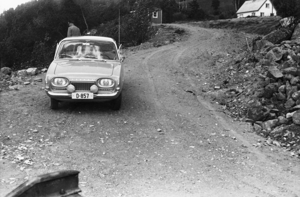 En Ford Taunus 17M av den typen som ble produsert i perioden 1960-64, fotografert forfra på den lille vegstubben som førte ned til den nye velteplassen for tømmer som var bygd for utislag av buntet tømmer fra lastebiler i Mjøsa ved Freng på Brøttum. Personbilen var D-registrert og hadde følgelig en tilhørighet til Hedmark fylke. En mann, antakelig en fløtingsfunksjonær som var på befaring, sto bak bilen. Høyere oppe i vegen ser vi ei Moelven-brakke, og helt til høyre øverst et kvitmalt bolighus. Bildet ble tatt i 1961, da den nevnte velteplassen var helt nybygd.