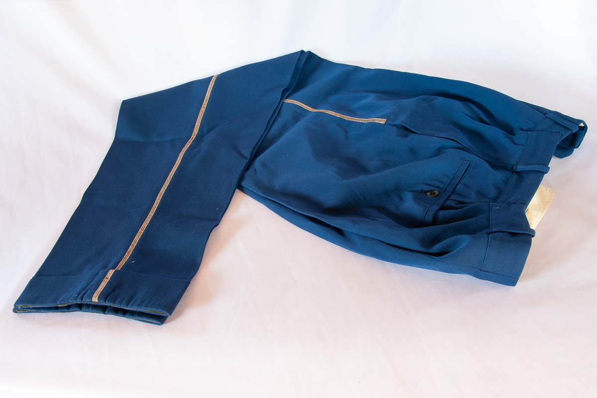 Uniformsbukse. Blå gabardin. Gullig pyntebånd langs buksebeina. Oppbrett. Gylf lukkes med 6 sorte plastknapper. To baklommer med sorte knapper bak, to stikklommer foran.  Lommene består av gulhvitt bomullstoff. Samme stoff rundt linning og gylf. Linning gjort mindre med inntak i blått.