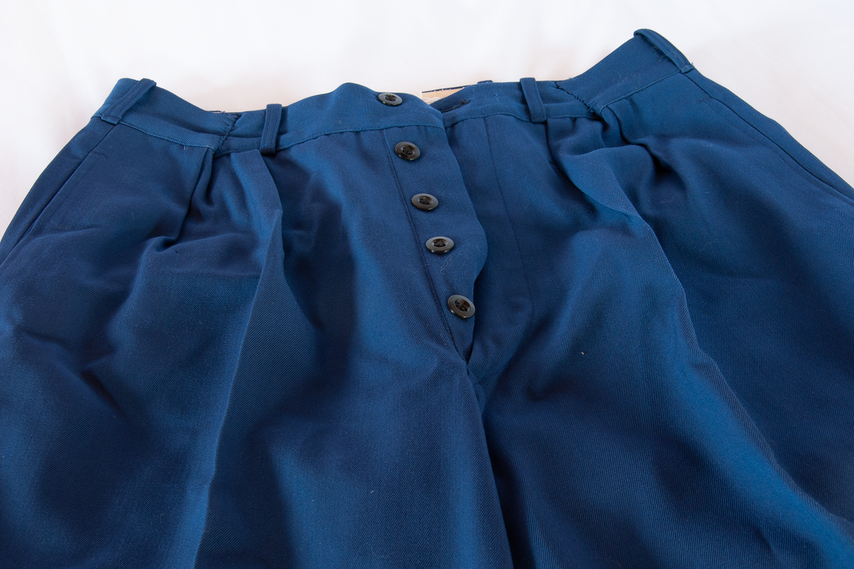 Uniformsbukse. Blå gabardin. Gullig pyntebånd langs buksebeina. Oppbrett. Gylf lukkes med 6 sorte plastknapper. To baklommer med sorte knapper bak, to stikklommer foran.  Lommene består av gulhvitt bomullstoff. Samme stoff rundt linning og gylf. Linning gjort mindre med inntak i blått.