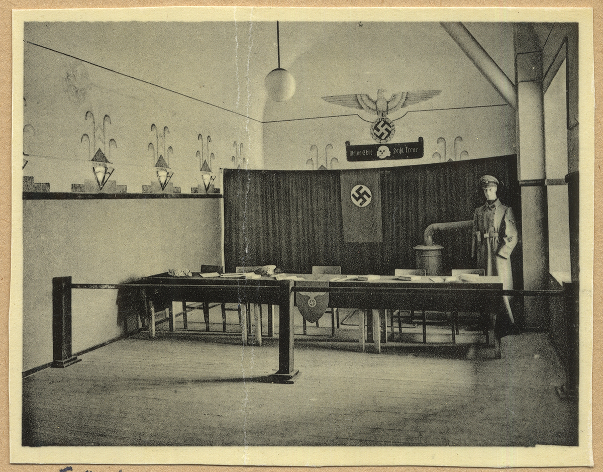 Bilden visar förhörssalen i koncentrationslägret Fort Breendonk i Belgien. Bakom en stängsel står en rad bord med stolar. Rummet är smyckat med nazistiska symboler. I hörnet står en docka med uniform.