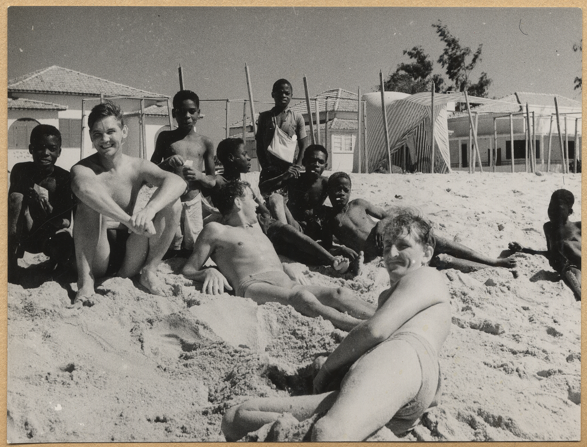 Svenska sjömän ligger på en sandstrand vid Afrikas kust omringat av svarta barn.