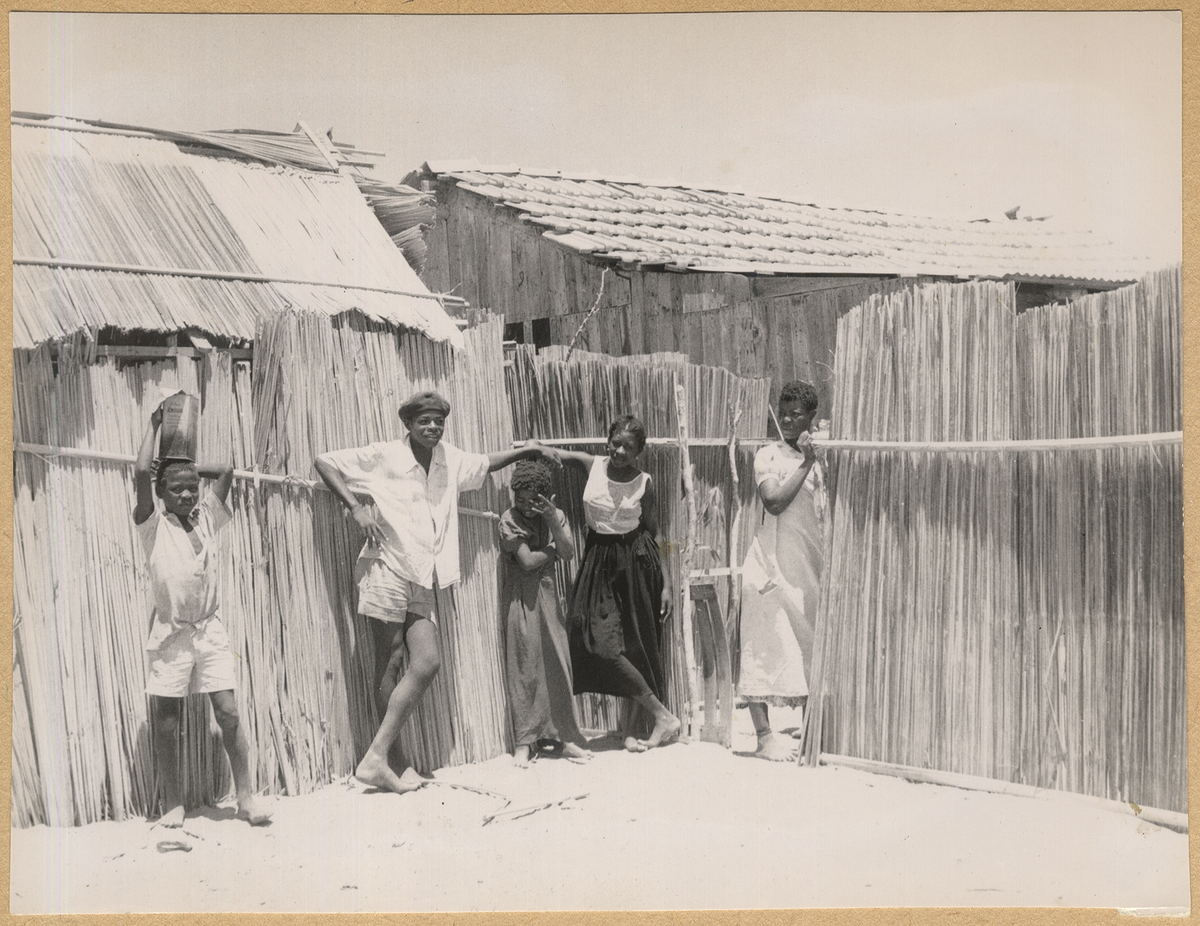Bilden visar en afrikans familj som står vid inhägnaden för sina enkla trähus i bakgrunden.