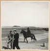 Två sjömän från Gotland promenerar på stranden. I bakgrund rider en svart man på en häst.