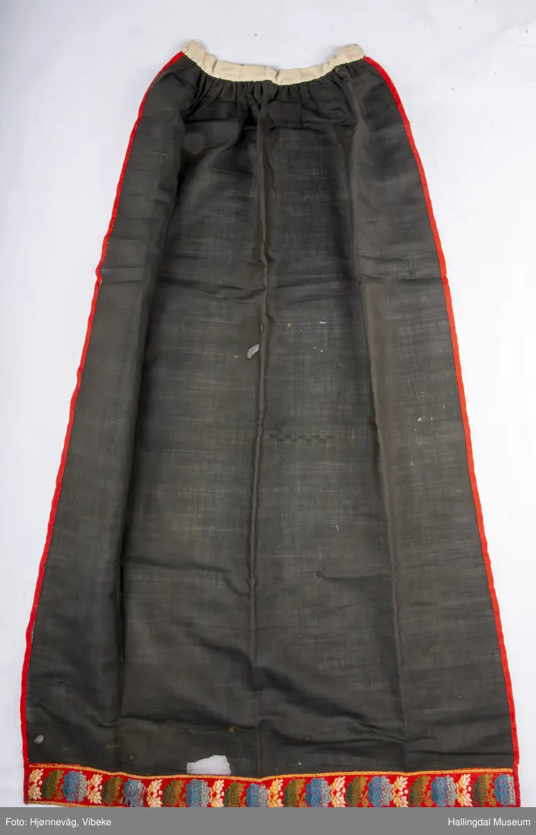 Fyriklæ i bomull med fløyelsband og ullband. Svart, tunt tøy i fyriklæet, liknar noko på bomull, men er noko stivare. Toskaft-veving. Rynka øvst, kvit bomullslist sett på øvst, br. 2,4 cm. Over denne festa ei bolu (svart fløyelsband med blomemønster i fiolett), l.: 38 cm og br.: 4 cm. Nedst eit tunt ullband, br.: 5,8 cm, blomemønster i 3 blåfargar, 3 grønfargar, kvitt og gult på raud botn. Langs sidene raudt ullband, 0,7 cm. Fyriklæet er sundt og bøtt sumstand.