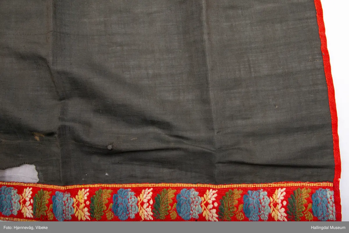 Fyriklæ i bomull med fløyelsband og ullband. Svart, tunt tøy i fyriklæet, liknar noko på bomull, men er noko stivare. Toskaft-veving. Rynka øvst, kvit bomullslist sett på øvst, br. 2,4 cm. Over denne festa ei bolu (svart fløyelsband med blomemønster i fiolett), l.: 38 cm og br.: 4 cm. Nedst eit tunt ullband, br.: 5,8 cm, blomemønster i 3 blåfargar, 3 grønfargar, kvitt og gult på raud botn. Langs sidene raudt ullband, 0,7 cm. Fyriklæet er sundt og bøtt sumstand.