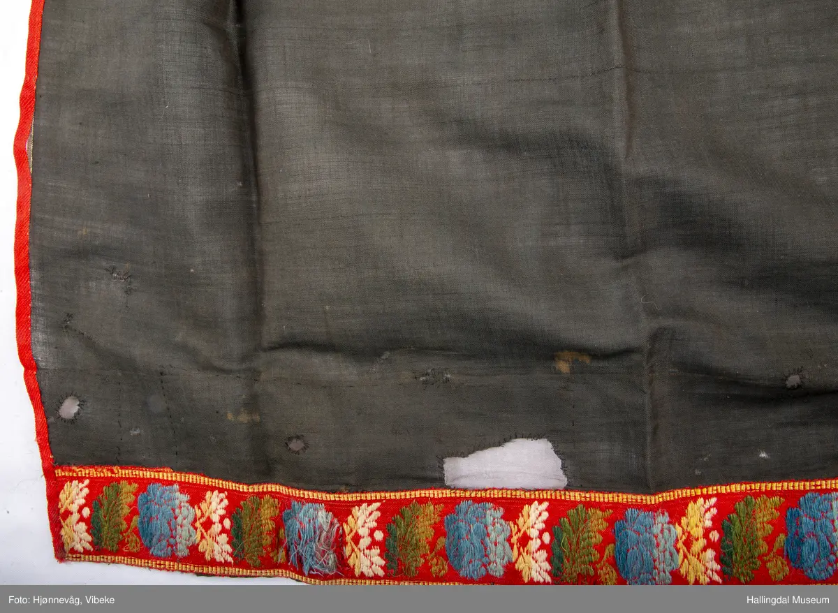 Fyriklæ i bomull med fløyelsband og ullband. Svart, tunt tøy i fyriklæet, liknar noko på bomull, men er noko stivare. Toskaft-veving. Rynka øvst, kvit bomullslist sett på øvst, br. 2,4 cm. Over denne festa ei bolu (svart fløyelsband med blomemønster i fiolett), l.: 38 cm og br.: 4 cm. Nedst eit tunt ullband, br.: 5,8 cm, blomemønster i 3 blåfargar, 3 grønfargar, kvitt og gult på raud botn. Langs sidene raudt ullband, 0,7 cm. Fyriklæet er sundt og bøtt sumstand.