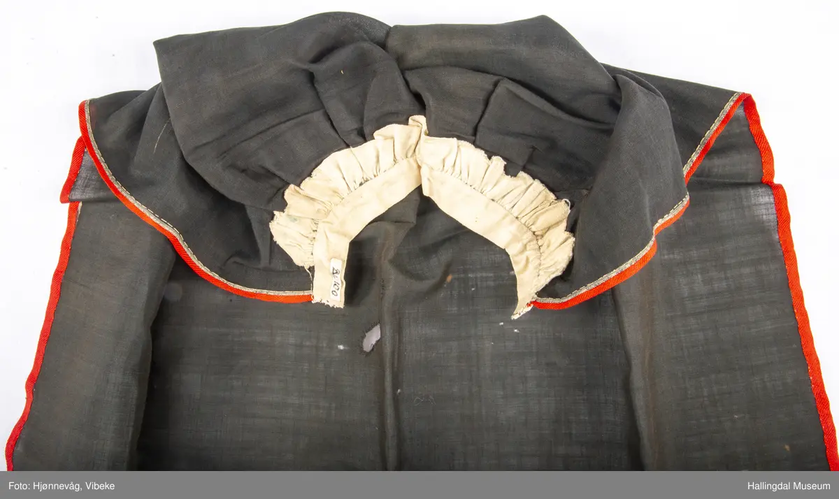 Fyriklæ i bomull med fløyelsband og ullband. Svart, tunt tøy i fyriklæet, liknar noko på bomull, men er noko stivare. Toskaft-veving. Rynka øvst, kvit bomullslist sett på øvst, br. 2,4 cm. Over denne festa ei bolu (svart fløyelsband med blomemønster i fiolett), l.: 38 cm og br.: 4 cm. Nedst eit tunt ullband, br.: 5,8 cm, blomemønster i 3 blåfargar, 3 grønfargar, kvitt og gult på raud botn. Langs sidene raudt ullband, 0,7 cm. Fyriklæet er sundt og bøtt sumstand.