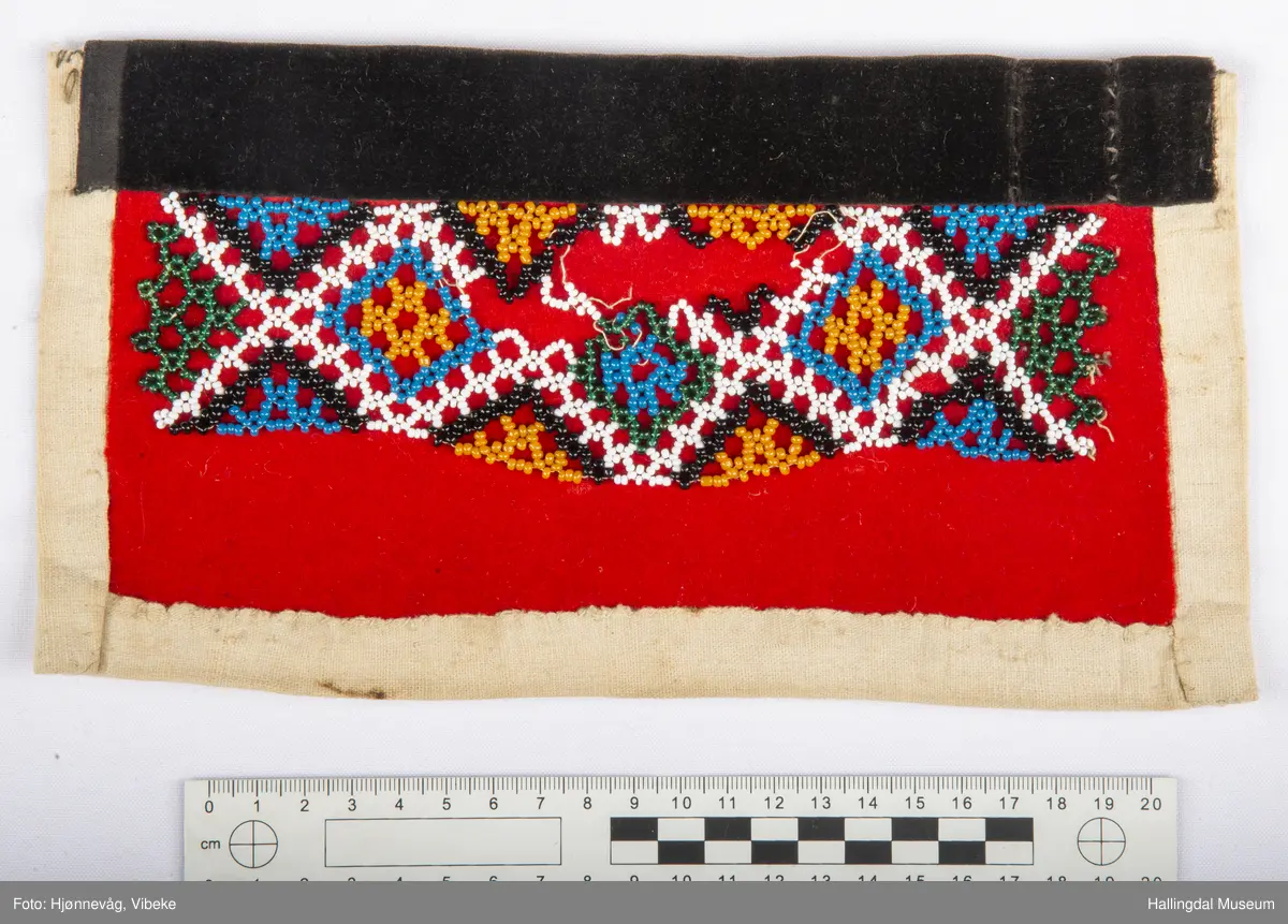 Kvit bomullslerret på baksida og bretta noko over på framsida. På frramsida raud vadmål med påsydd geometrisk mønster av små, runde glasperler. Øvst på brystduken ein svart fløyelskant.
