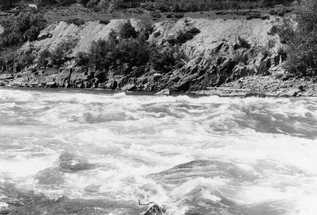 Fra nedre del av Gudbrandsdalslågen, nærmere bestemt strekningen mellom Hunderfossen og Isakstuøra, der elva var kommunegrense mellom Øyer på østsida og Fåberg på vestsida av vassdraget. Dette fotografiet er tatt fra elvebrinken på Øyer-sida, med kameraet vendt skrått i medstrøms retning, på et sted der Lågen rant gjennom et berglendt terreng med et visst fall. På Fåberg-sida, øverst til venstre i bildet, skimter vi et parti der det sto ei pålerekke. Dette fotografiet ble tatt i 1961, altså på et tidspunkt da representantskapet i selskapet Opplandskraft hadde kjøpt fallrettighetene i Hunderfossen og Ensbyfallene med sikte på kraftutbygging og startet forbedende prosjekteringsarbeider. Ettersom gjeldende lov om vassdragsreguleringer forutsatte at regulanten skulle tilstrebe at inngrepene i minst mulig grad skulle medføre ulempe og skade på eksisterende bruk av vassdragene, er det nærliggende å anta at fløtingsfunksjonæren som tok dette fotografiet var opptatt av å dokumentere forholdene før kraftverket ble etablert. På det albumarket der kopien av dette fotografiet ble montert finner vi følgende notat fra ei befaring Dette fotografiet er tatt under ei befaring 25. juni 1961, da vannføringa i Losna (noe høyere oppe i elveløpet) var 252 kubikkmeter i sekundet. Dette bildet er tatt fra østre elvebredd, på tvers av elveløpet, som her gikk kvitskummende i et åpenbart steinete lende, som nok ville skapt problemer for tømmerfløterne om vannføringa hadde vært mindre. Som en kommentar til akkurat dette motivet har fotografen skrevet:

«Bilde nr. 40, det samme i enda sterkere stryk.» Med «det samme» refereres til omtalen av tidligere bilder i samme serie, som lyder slik: «7. Sammenhengende strykparti i ca. 1 000 m. lengde til ovenfor Hunderfoss jernbanebro. En anselig mengde grov stein synlig i fossens djupål eller kan skimtes i fossestryket. Ved ytterligere red. av vannføringen vil antall steiner farlig for fløtningen øke sterkt. En steinør av betydelig omfang strekker seg langs østre land nedenfor jernbanebroen. Det er ikke noe tømmer ute i stryket, men langs land, og spesielt på øra, ligger en god del. Bilde nr. 32 og 34 gir et inntrykk av den grovkuperte elvebunn.»

For Glomma fellesfløtingsforenings del var det knyttet atskillig spenning til hvordan fløtinga ville arte seg på denne strekningen når kraftstasjonen ved Hunderfossen ble satt i drift. Fra da av ville nemlig mye av vannet gå gjennom turbinene og via en underjordisk avløpskanal som skulle munne ut i Lågen igjen ved Isakstua, snaut fire kilometer nedenfor.