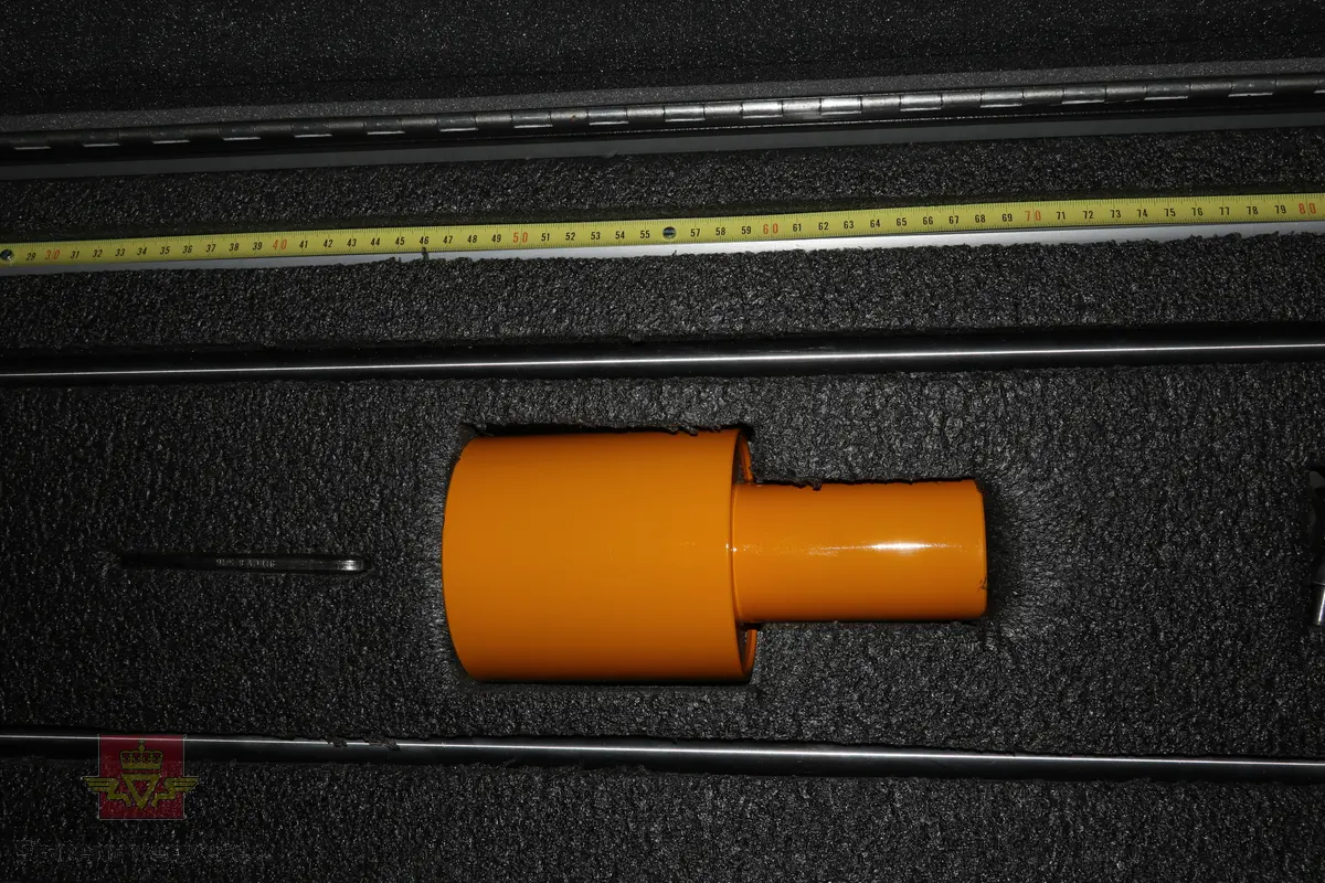 Sort trekasse med bærehåndtak som inneholder et mekanisk målemeter av stål som består av flere bevegelige deler som en sleidehammer med måleskala. I bunnen av utstyret sitter en utskiftbar metallspiss. Kofferten inneholder 40stk ekstra metallspisser.