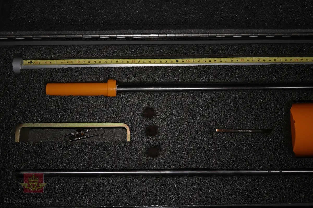 Sort trekasse med bærehåndtak som inneholder et mekanisk målemeter av stål som består av flere bevegelige deler som en sleidehammer med måleskala. I bunnen av utstyret sitter en utskiftbar metallspiss. Kofferten inneholder 40stk ekstra metallspisser.